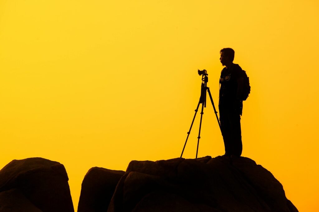 Silhouette of a lone photographer with tripod on a rocky cliff against a golden sunset sky.